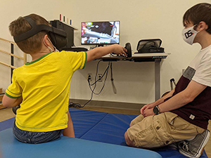 Child wearing VR headset uses one handheld controller to reach in a video game merging therapy and fun; a research assistant observes.