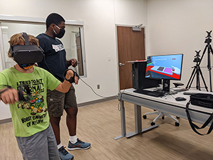 Child wearing VR headset uses handheld controller to reach in a video game merging therapy and fun; an undergraduate research assistant observes.