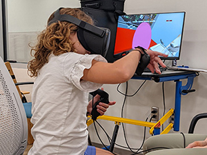 Child wearing VR headset coordinates two handheld controllers to scoop ice cream in a video game merging therapy and fun; a research assistant observes.