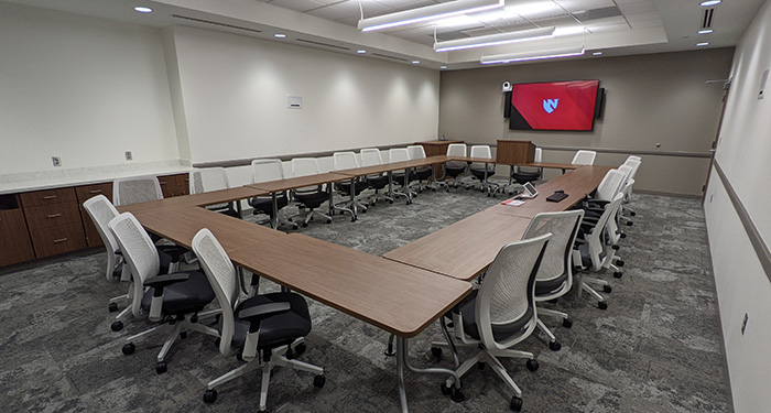 Medium conference room, MMI 40032, tables rectangular formation with 24 chairs on outer edge, portable tabletop podium, computer, webcam, keyboard, mouse, 2 large flat screen TVs on opposite ends of room.