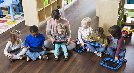 stock photo of early childhood teacher sitting on the floor with children working on electronic devices together; credit: iStock
