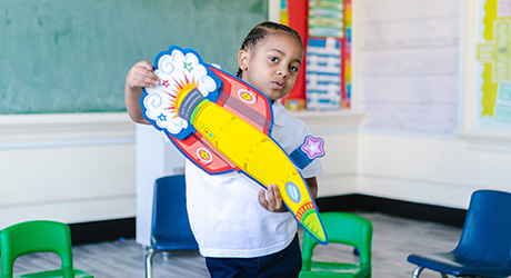 school aged boy in black pants and white t-shirt in a classroom holding a colorful paper spaceship; credit Rodnae Productions from Pexels