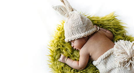 Baby in white knit bunny ears cap, laying on belly on green fur; Credit Gigin Krishnan from Unsplash