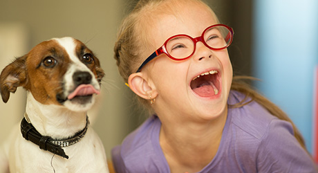 stock photo of young girl with developmental disability, wearing glasses, laughing, big smile, next to jack russell terrier dog sticking out its tongue; credit Shutterstock