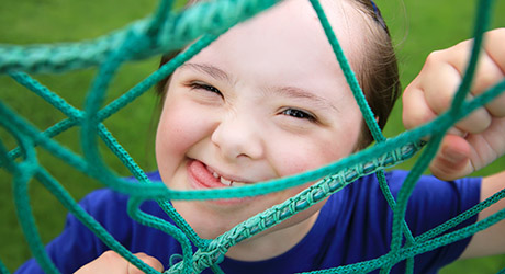 stock photo of young child playing outdoors, climbing on a net ladder, squinting eyes, tongue out through a smile; credit: Shutterstock