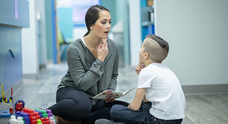A speech-language pathologist and child sitting on the floor working together to form speech sounds with their mouths, one is holding a book; credit iStock.