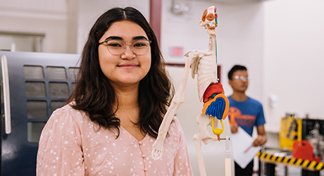 Student with dark hair below shoulders, wearing eye glasses and long-sleeve pink shirt with white stars, standing beside human body skeleton model; another student in background is out of focus; credit Jeswin Thomas from Unsplash.