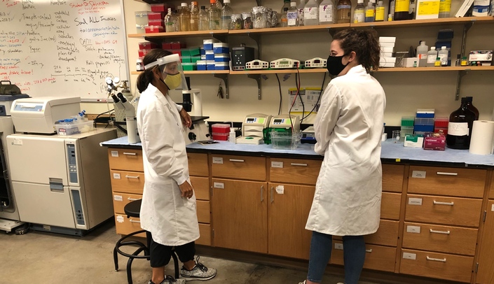 Dr. Annemarie Shibata, at left, talks with undergraduate research student, Carly Baker, in her lab at Creighton University, while adhering to social distancing and other COVID-19 CDC recommended safety guidelines.