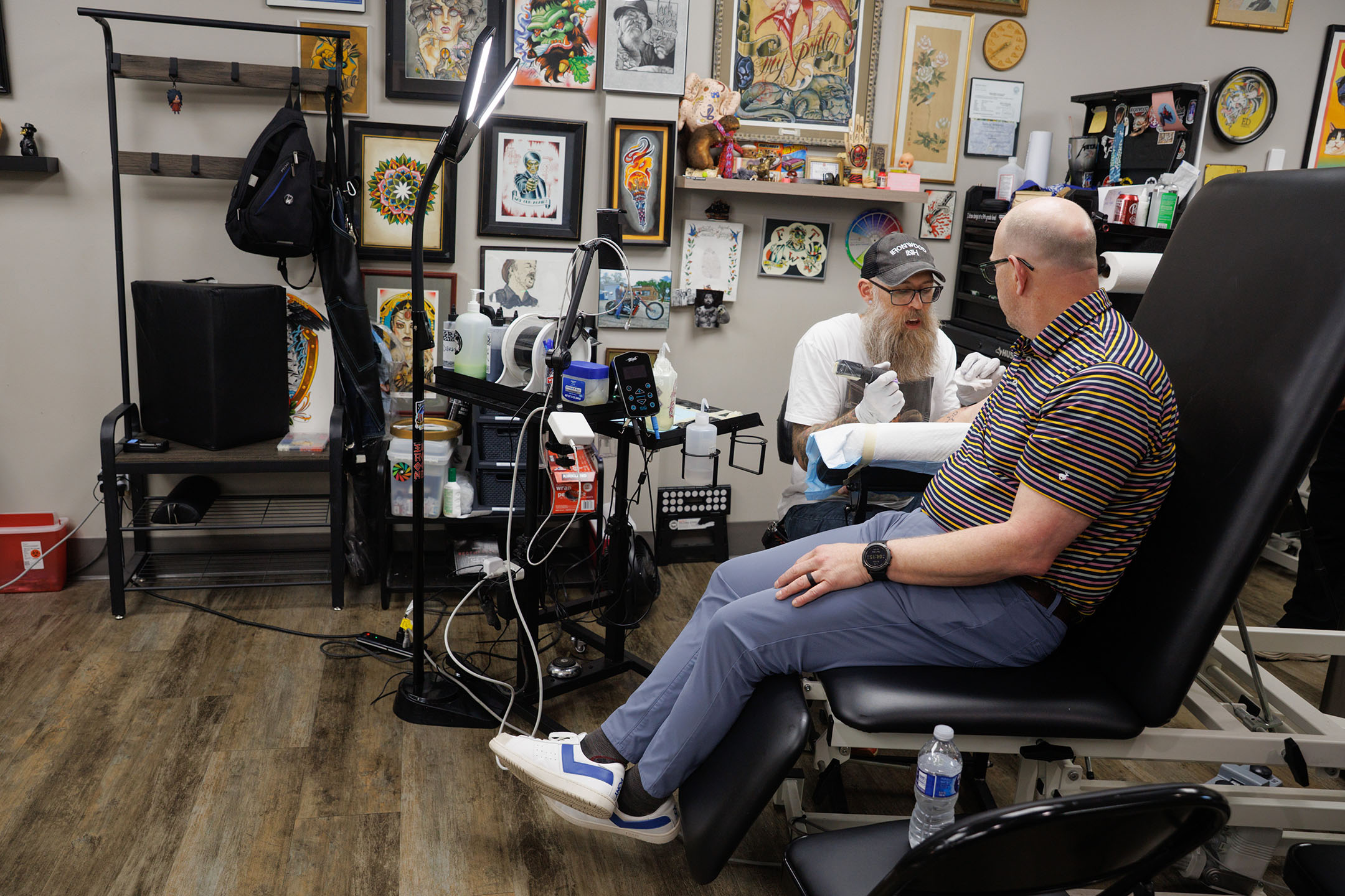 Corey Hopkins, PhD, sitting on a chair at a tattoo parlor where a tattoo artist is at work
