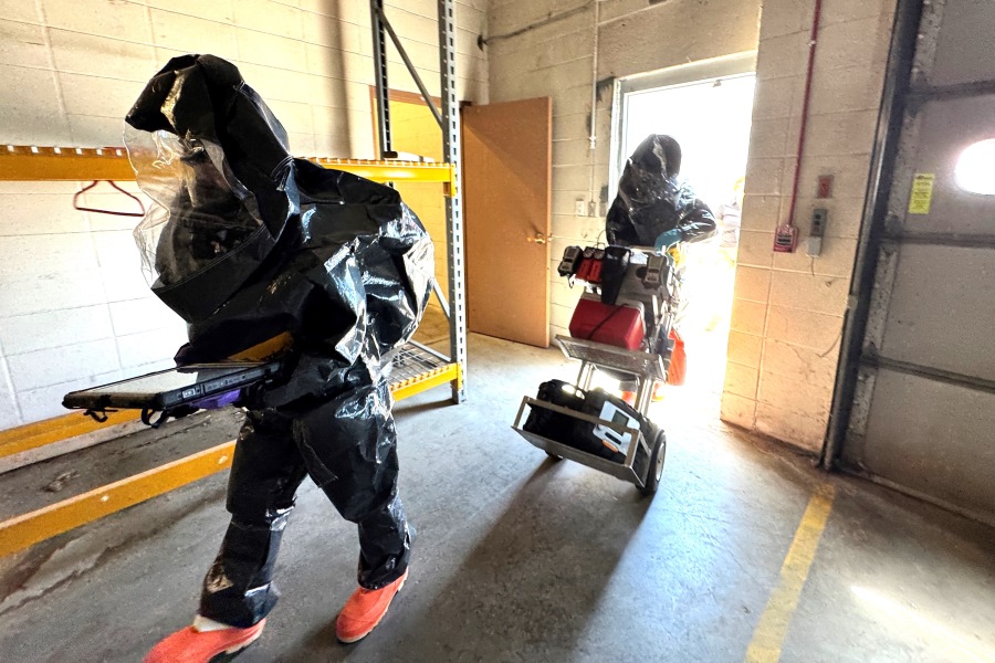 Two people in protective suits enter building with gear