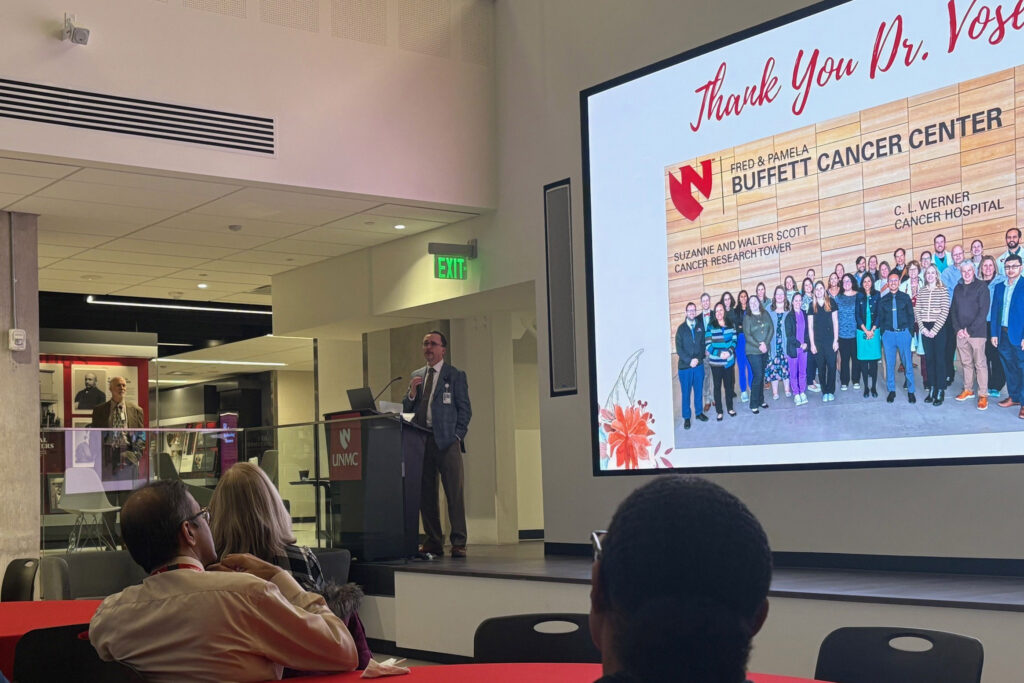 Dr. Michael Ash, in a business suit, speak at a podium in front of a crowd at a December event celebrating Julie Vose, MD. 