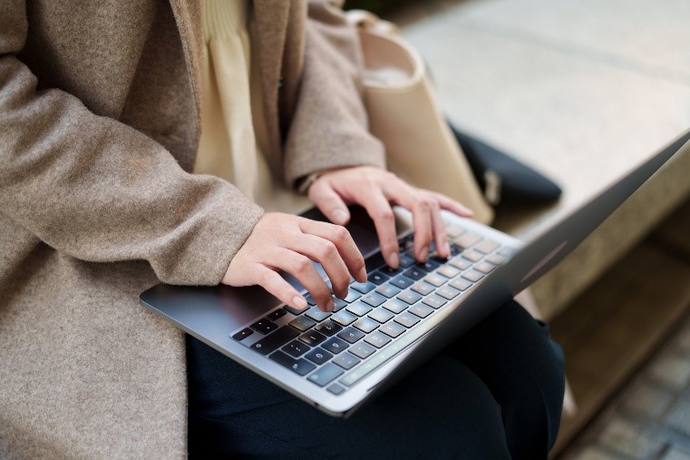 Photograph of a person typing on a laptop keyboard while seated outdoors.