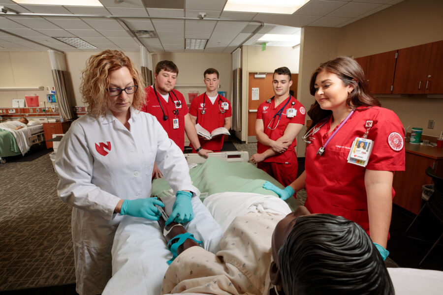 A nursing instructor administers an injection to a medical manekin while four students stand and watch.