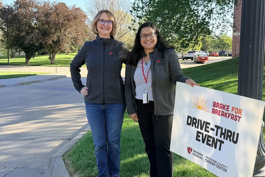 Two women stand outdoors next to "Brake for Breakfast Drive-Thru Event." The sign features a sun graphic, event title in bold letters, and logos for Nebraska Medicine and Fred & Pamela Buffett Cancer Center.