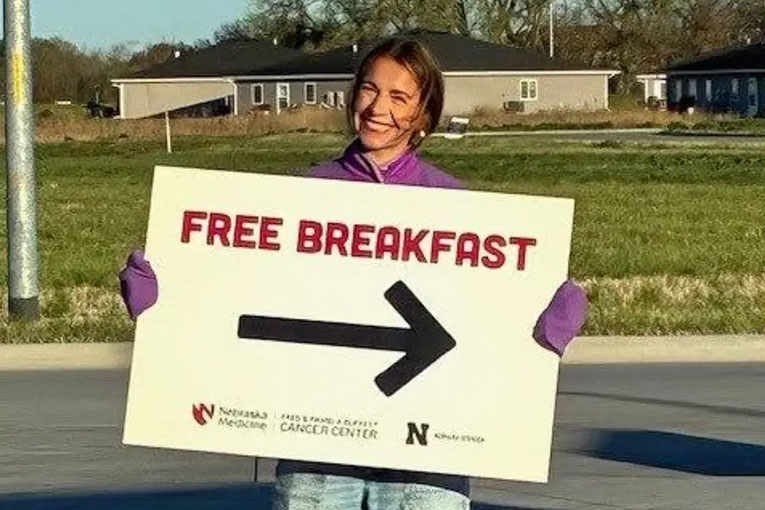 Photograph of a person holding a large sign with bold red text "FREE BREAKFAST" and a black right-pointing arrow. Sign includes logos for Nebraska Medicine, Fred & Pamela Buffett Cancer Center, and Nebraska.
