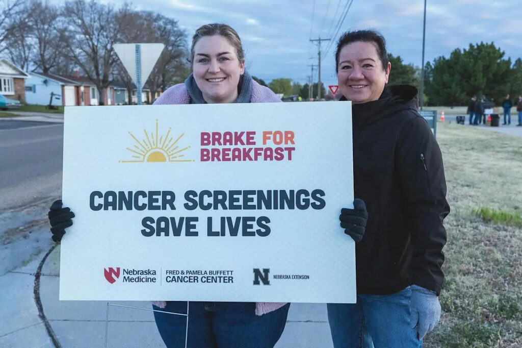 Photograph of two people holding a sign promoting cancer screenings with text "Brake for Breakfast" and "Cancer Screenings Save Lives," along with logos for Nebraska Medicine, Fred & Pamela Buffett Cancer Center, and Nebraska Extension.
