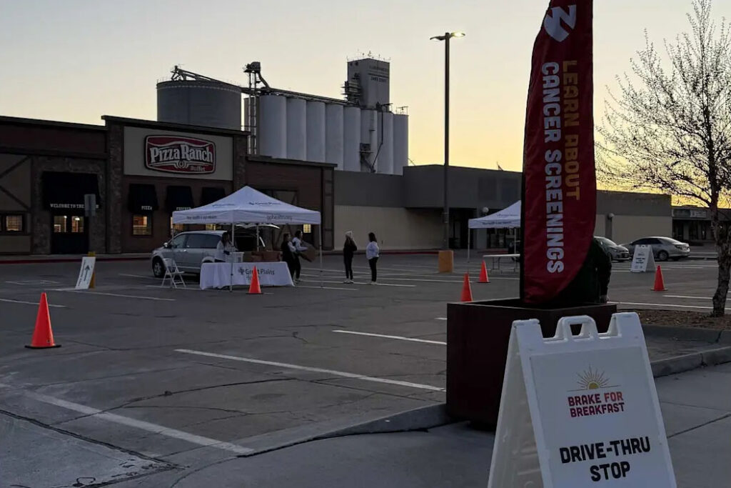 Photograph of a Brake for Breakfast drive-thru event promoting cancer screenings, held in a parking lot near a Pizza Ranch restaurant at sunrise. A tall red banner says, "Learn About Cancer Screenings."
