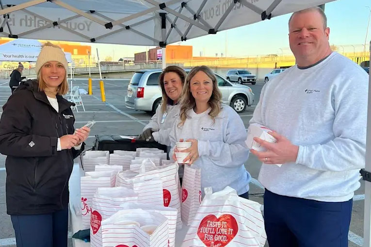 Photograph of four people standing behind a table filled with stacked Chick-fil-A food bags under a tent in a parking lot.