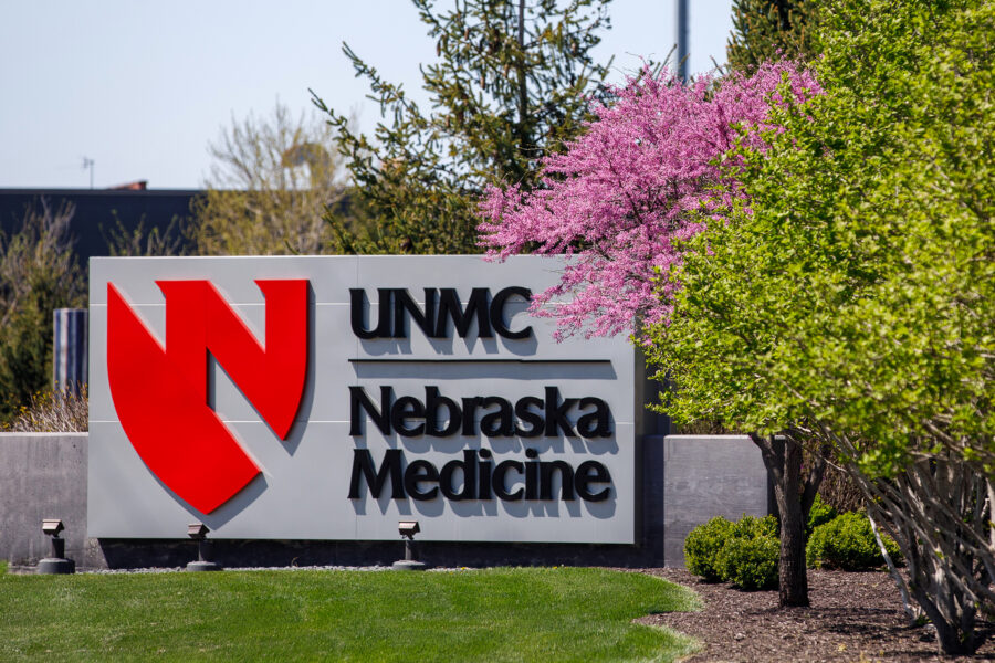 A med center entrance sign that has the words UNMC | Nebraska Medicine and their shared shield logo with blooming trees in the foreground of the sign.