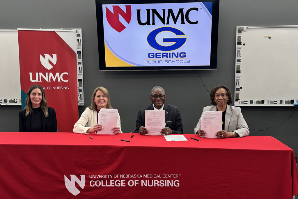 Attending the MOU signing ceremony were, from left, Haley Hays, DNP, interim assistant dean of the UNMC College of Nursing-Western Division; Superintendent Nicole Regan, EdD, of Gering Public Schools; UNMC Interim Chancellor H. Dele Davies, MD; and Lepaine Sharp-McHenry, DNP, dean of the UNMC College of Nursing. They are seated at a table holding the agreement with the logo for UNMC on the tablecloth and the logo for UNMC and Gering Public Schools on a screen behind them.