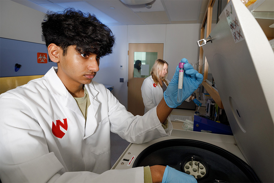 Vamsi Singu, participating in the UNMC Department of Anesthesiology's Summer Training in Anesthesia Research (STAR) program, working in a lab while wearing a UNMC lab coat and holding up a labeled beaker, with a lab researcher in the background.