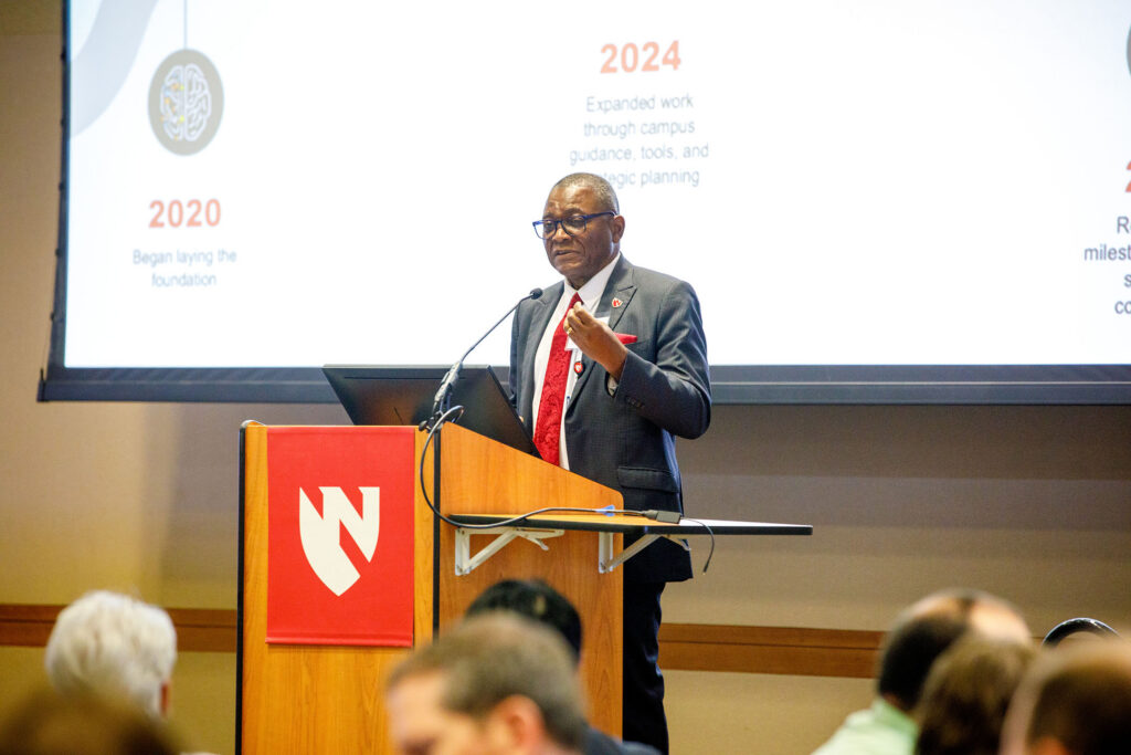 UNMC Interim Chancellor H. Dele Davies, MD, gives introductory remarks at the "AI in Health Conference." He is speaking from behind a podium with the UNMC/Nebraska Medicine shield logo and a drop down screen display behind him.