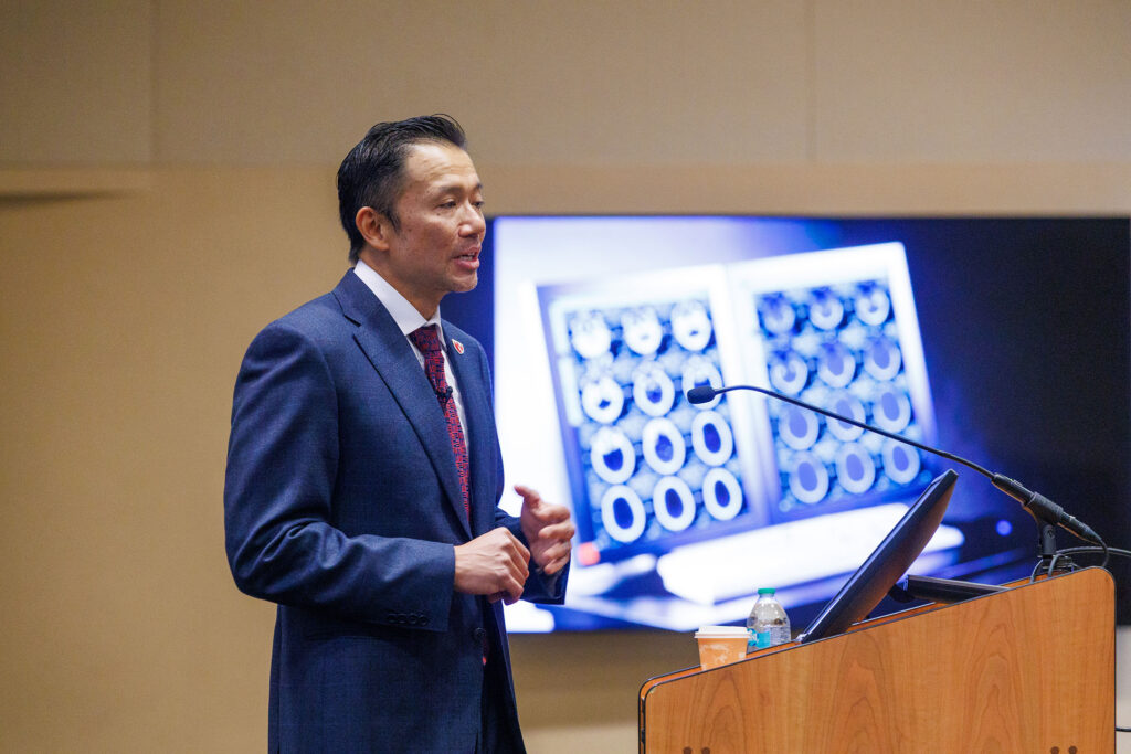 Ryan Lee, MD, chair of the UNMC Department of Radiology, gives the keynote speech at UNMC's "AI in Health Conference 2026: Shaping the Future of Care." He is speaking from behind a podium with an image of medical scans behind him in the frame.