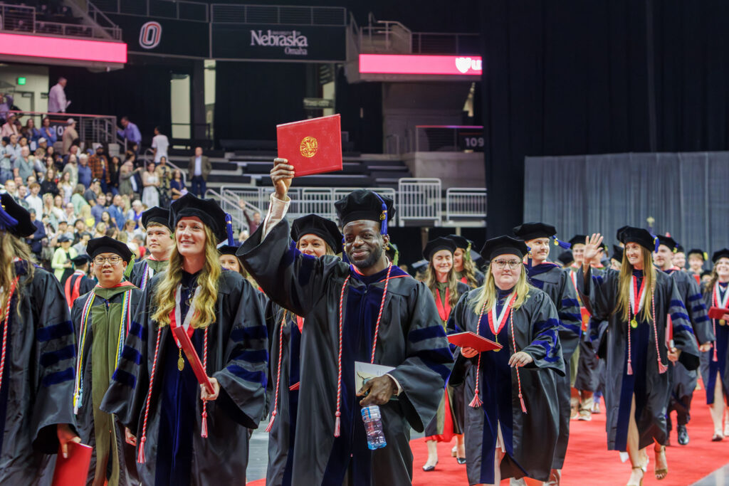 As graduates depart, a UNMC graduate holds up his degree and smiles.