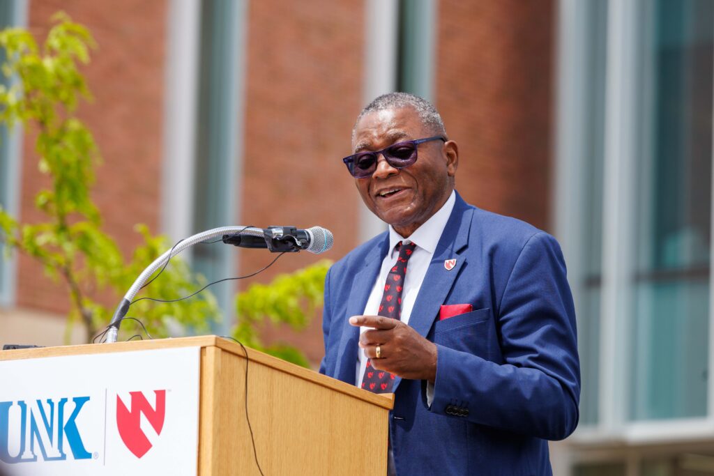 UNMC Interim Chancellor H. Dele Davies, MD, in dark glasses and a blue jacket, points forward with a smile as he speaks from behind a microphone, standing at a podium with the dual logos of the University of Nebraska at Kearney and UNMC, at the formal opening of the Douglas A. Kristensen Rural Health Education Complex, including the new Health Science Education Center II, on May 14.