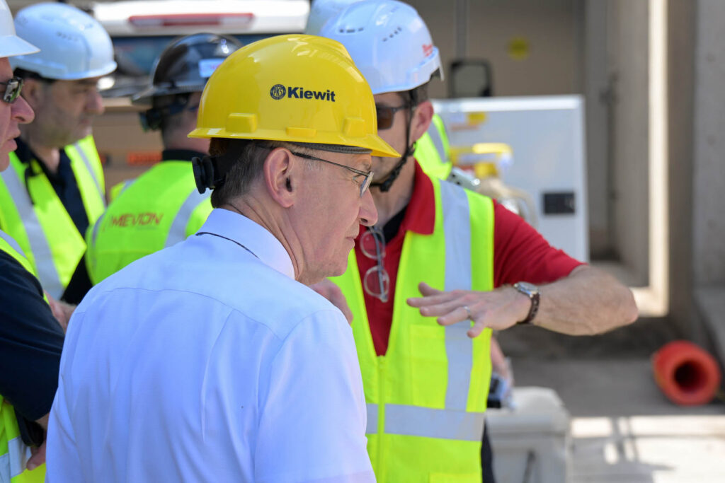 Dr. Charles Enke, wearing a yellow hard hat, looks to the right at the (unseen) system, as a worker in a white hard hat and yellow safety vest appears to be explaining something to Enke and a co-worker.