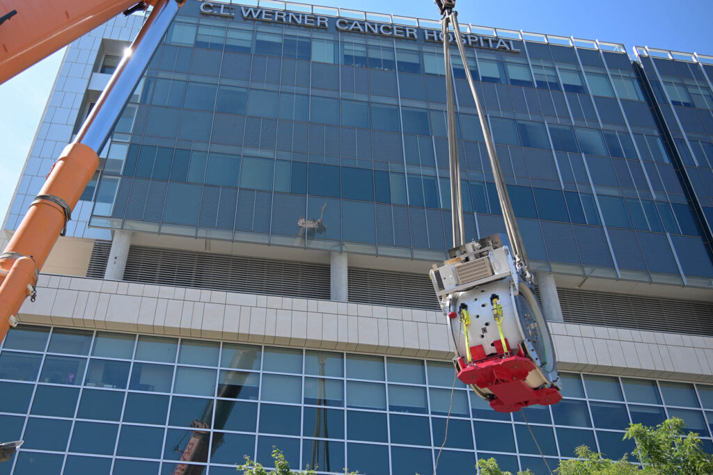 The proton beam therapy system, on an orange platform, is hoisted into the air by a crane in front of the CL Werner Cancer Hospital building, part of the Fred & Pamela Buffett Cancer Center.
