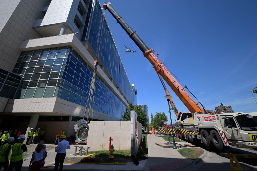 The roundish, futuristic looking proton therapy system, at the left of the photo, has been attached to a yellow crane at the right of the photo as workers and others look on in front of the Fred & Pamela Buffett Cancer Center.