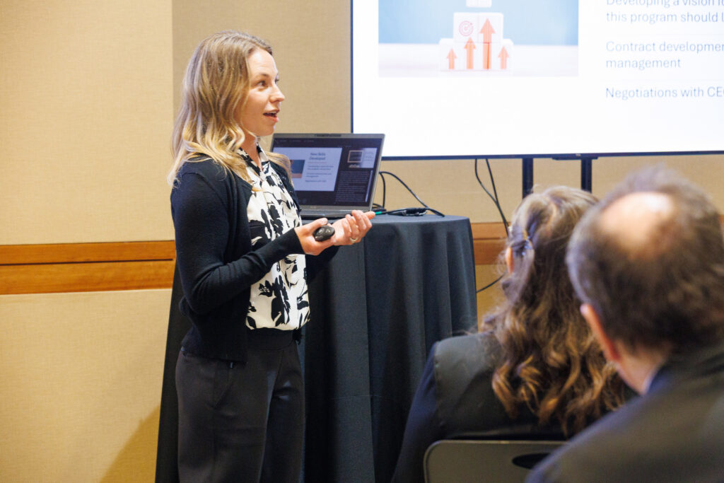 Rachel Knight, PhD, wearing a black jacket and a black-and-white patterned blouse, looks at the audience with her hands held palms up as she delivers her presentation.