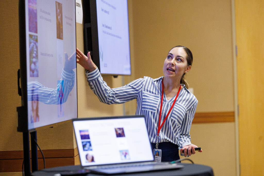 Chelsea Navarette, MD, in a striped blouse, looks at the screen and touches one side of it, with her laptop at the forground, as she delivers her presentation.