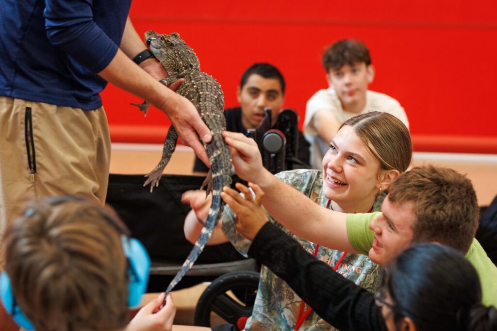 Kip Smith holds Tripod, a three-legged baby alligator, while smiling, seated campers reach forward to pet his body and tail.