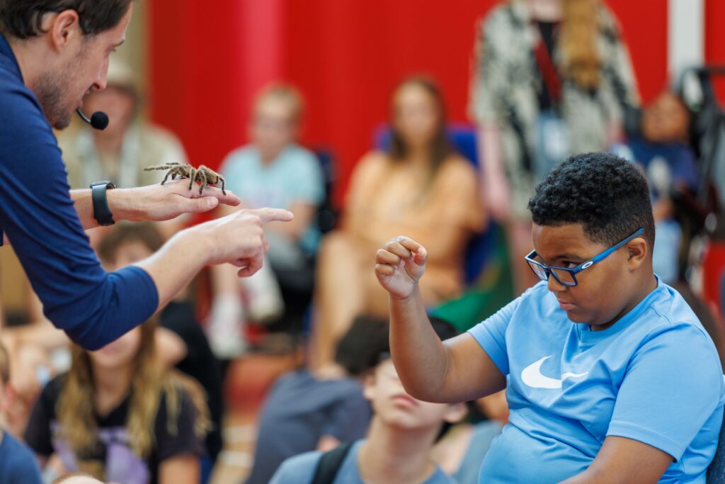 Kip Smith, left, displays a tarantula in his hand to camper Jahmir Blankman, seated at right in a blue Nike shirt and glasses. Blankman is reaching tentatively out toward the tarantula, but not touching it. 