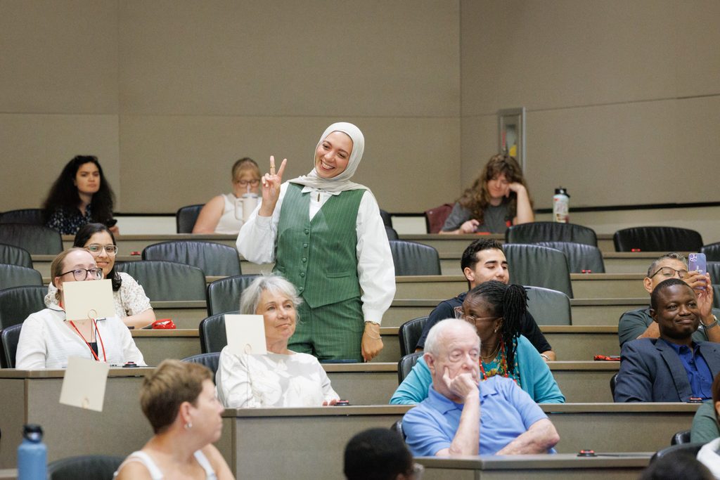 A student in green wearing hijab smiles and gives a peace sign to a roomful of other students, who are seated around her as she stands.