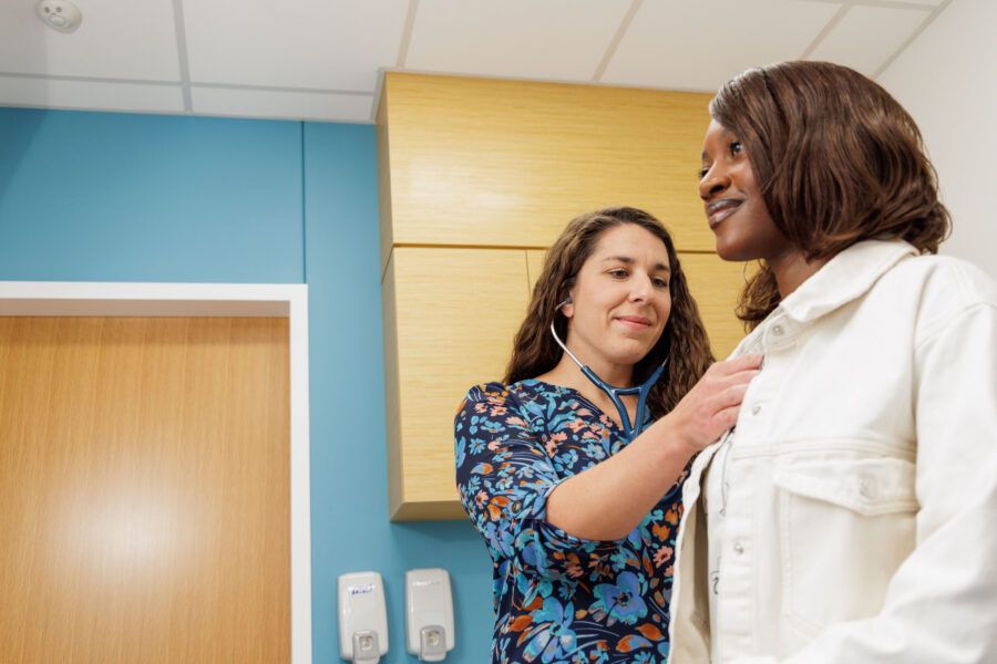 A health care provider in a blue floral shirt holds a stethoscope to the chest of a smiling young woman in a white shirt.