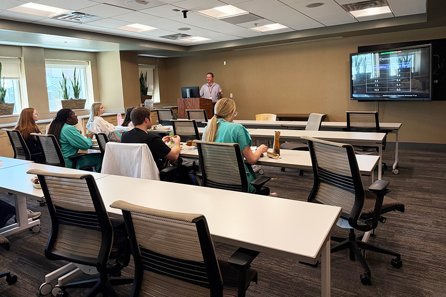 Classroom with male teacher standing at a podium in front, learners sitting in chairs, and a monitor on the front wall.
