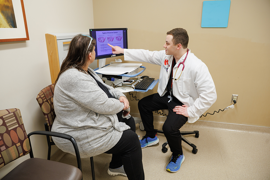 Patient sitting in a chair and a medical student wearing a white jacket, pointing to a computer screen.
