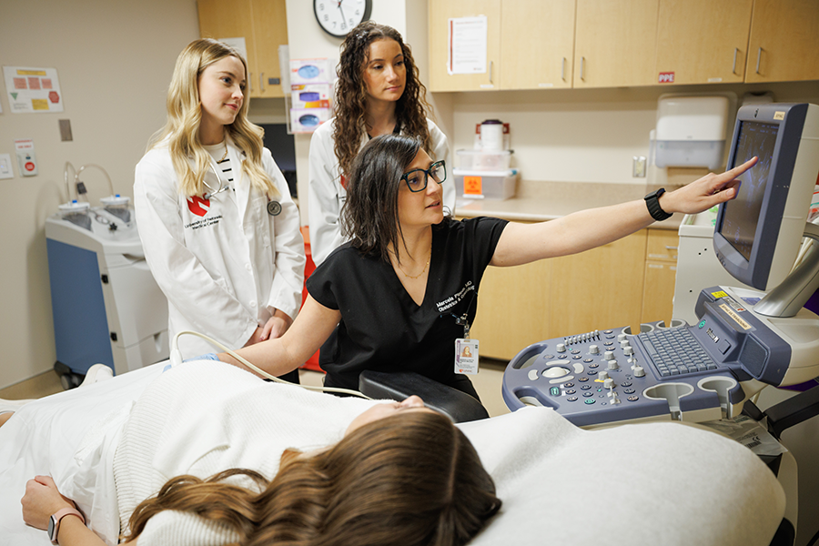 Physician performing an ultrasound on a pregnant patient, pointing to the screen, and two people wearing white coats observing.