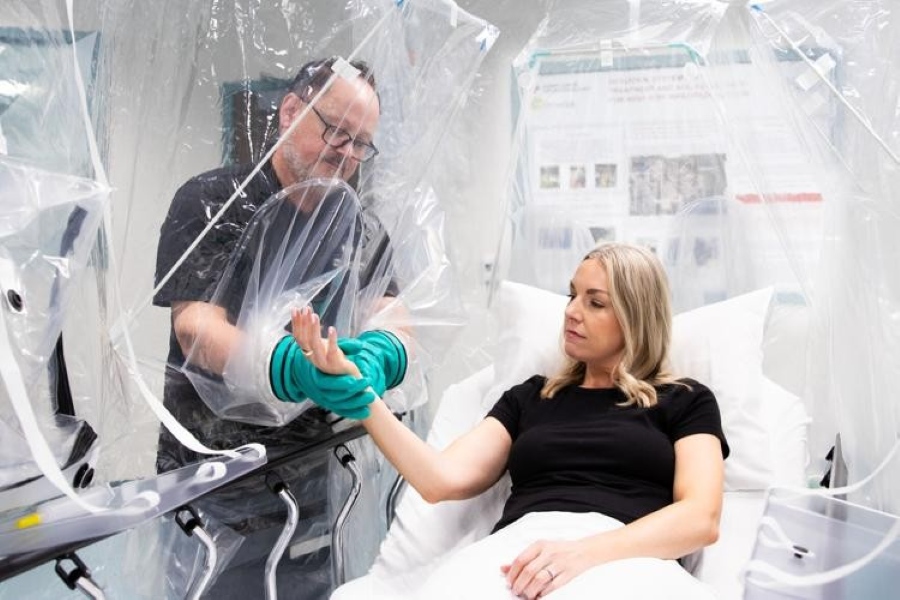 Woman in hospital bed enclosed in clear plastic is receiving care from man outside enclosure