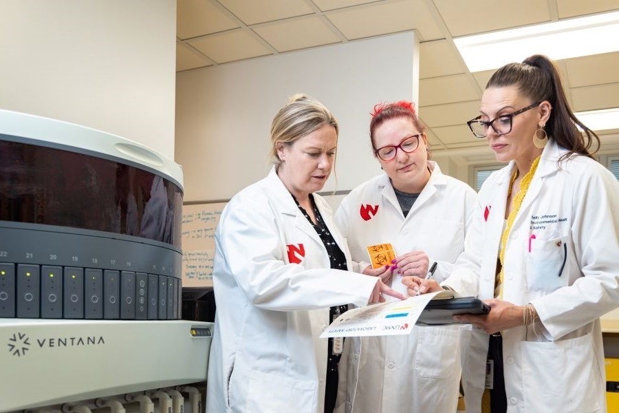 Three women in lab coats looking at clipboard in lab