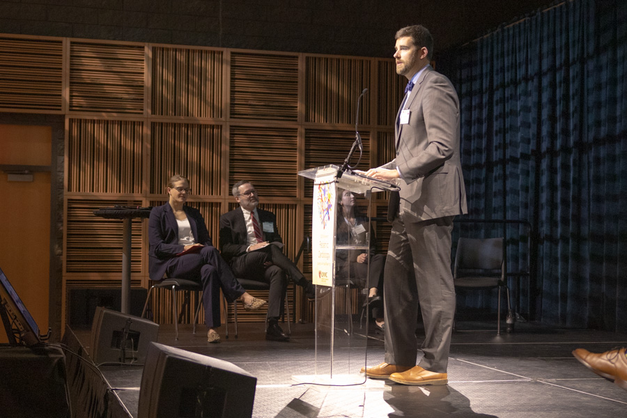 Man stands at podium with people sitting on stage in the background