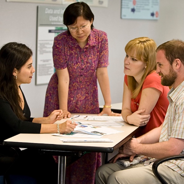 A professor instructing three students at their desks. 