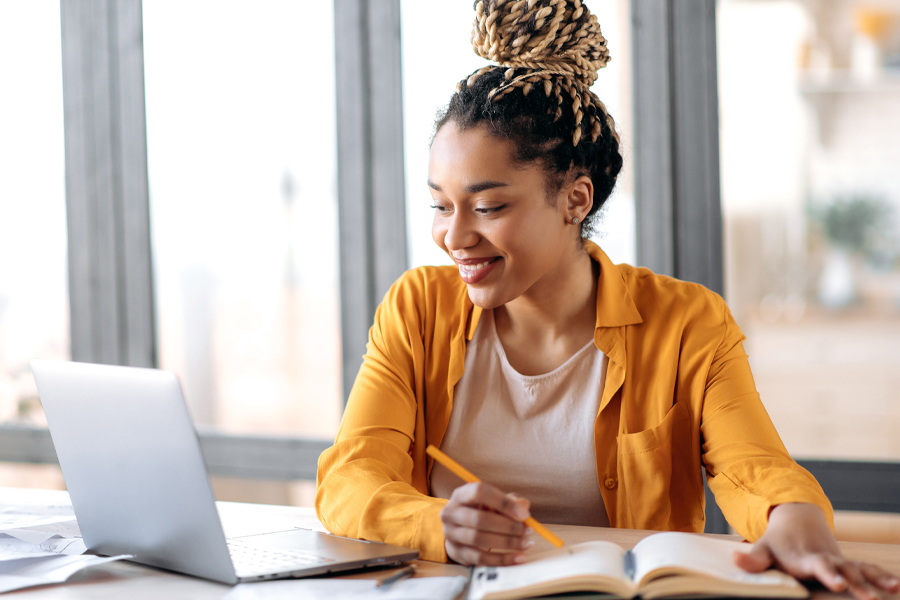 Woman sitting at table with laptop and notebook holding a pencil