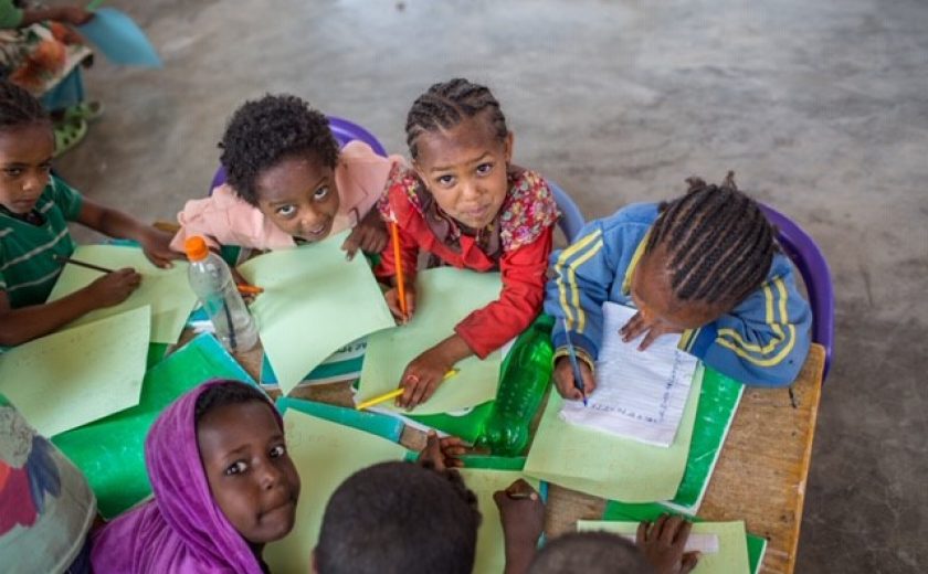 Image of children from Ethiopia sitting at a desk and writing on paper. Some students are looking at the camera.