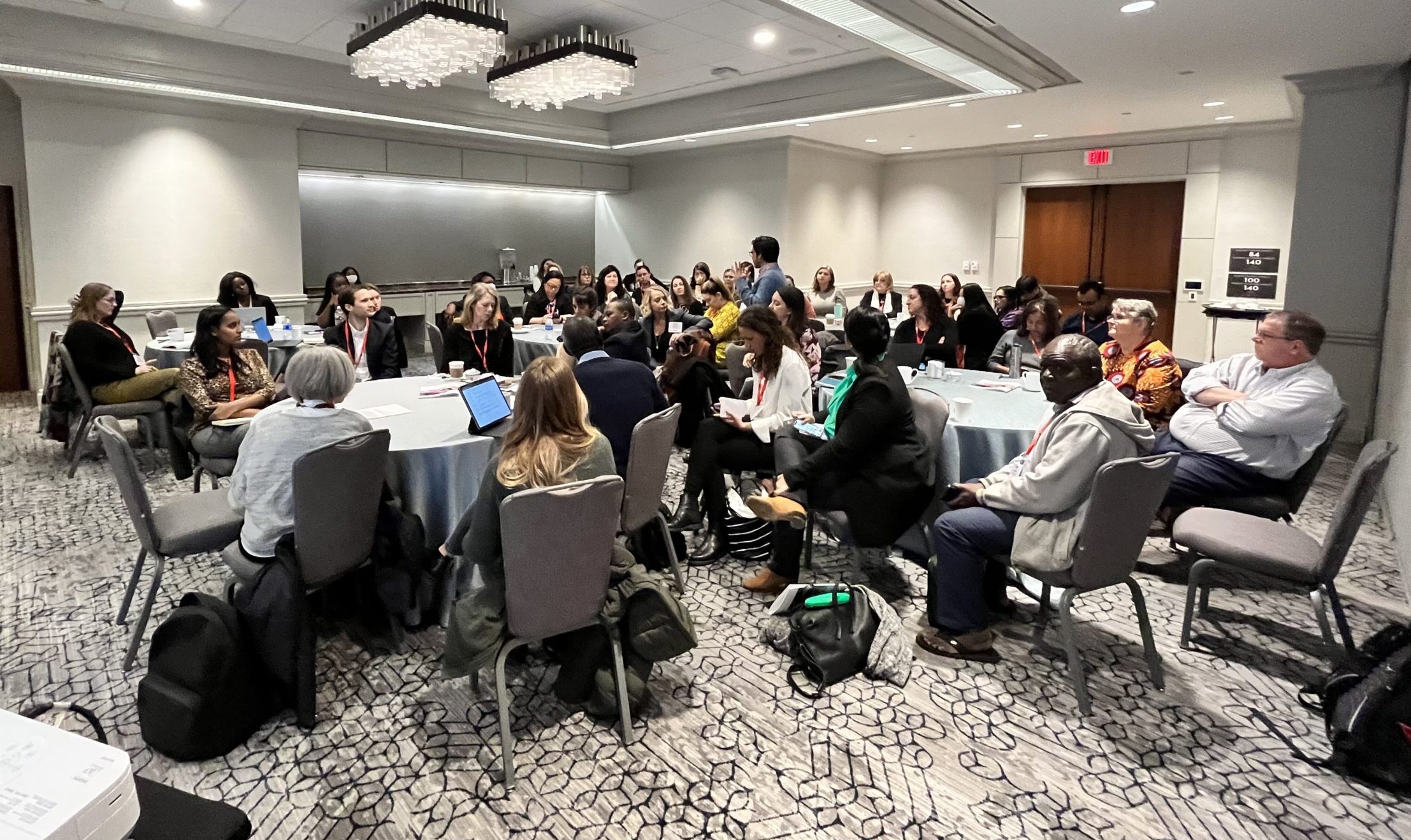 Image of meeting participants sitting around tables listening to a speaker.
