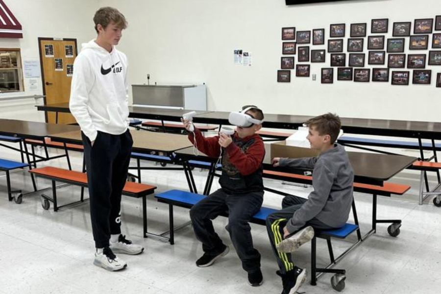 Three middle-school aged boys are in an empty school cafeteria, one wears a virtual reality headset while the others watch.