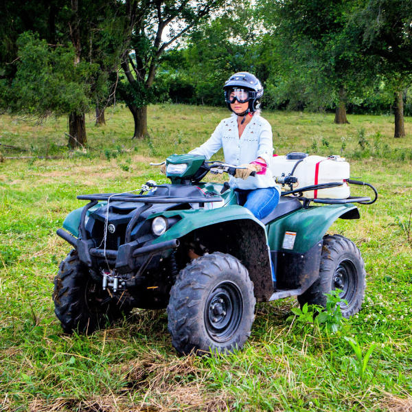 Woman riding an ATV with a sprayer mounted on the back in a grassy area. The woman is wearing a helmet, goggles, gloves, long pants, and long sleeves. 